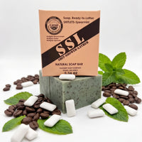 Front view of 'Flagship Soap Company' natural 'Shitlets-Spearmint' exfoliating soap bar (green with coffee specks, on a stack of bars) and its cardboard packaging box on a white background. The packaging displays the 'FLAGSHIP' seal, 'SHIT-SHOWER-LATHER', 'NATURAL SOAP BAR', and '5.56 OZ'. Surrounding the soap are piles of actual coffee beans, fresh green mint leaves, and several white spearmint gum chiclets, visually representing the spearmint and coffee natural exfoliants used in the soap's formula.