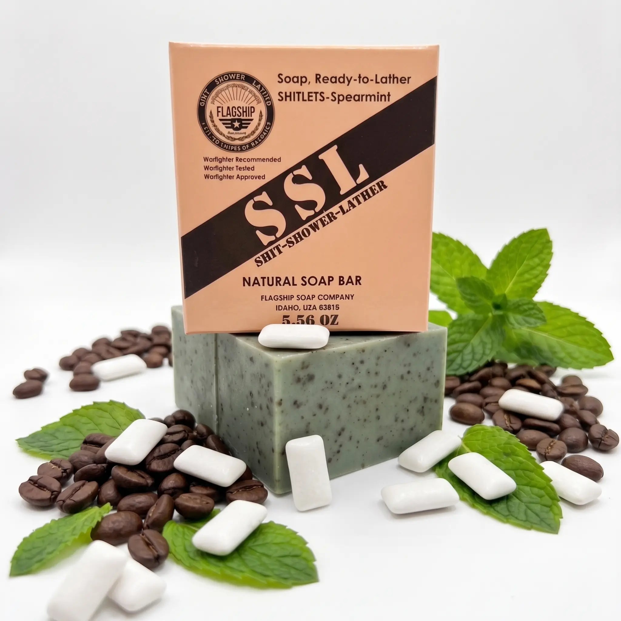 Front view of 'Flagship Soap Company' natural 'Shitlets-Spearmint' exfoliating soap bar (green with coffee specks, on a stack of bars) and its cardboard packaging box on a white background. The packaging displays the 'FLAGSHIP' seal, 'SHIT-SHOWER-LATHER', 'NATURAL SOAP BAR', and '5.56 OZ'. Surrounding the soap are piles of actual coffee beans, fresh green mint leaves, and several white spearmint gum chiclets, visually representing the spearmint and coffee natural exfoliants used in the soap's formula.