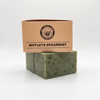 Front view of 'Flagship' Shitlets MRE-themed spearmint and coffee natural soap bar and its cardboard packaging box. The bar is green with exfoliating coffee specks and sea clay. The package clearly shows the 'Shit, Shower, Lather' slogan and the Flagship seal. High-performance, patriotic veteran gift soap from Flagship Soap Company.