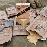 A flat-lay photograph featuring Holy Grail Natural Soap by Flagship Soap Company centered among various authentic Meals Ready to Eat (MRE) components. The soap bar and its tan "SSL" (Shit-Shower-Lather) themed packaging sit atop a "Meal, Cold Weather" packet, surrounded by an MRE cracker, beverage powder, and an open pouch of cheddar cheese sauce with jalapeños. Tactical gloves and a metal spoon are visible in the background on a green field jacket, emphasizing the rugged, veteran-inspired aesthetic. This g