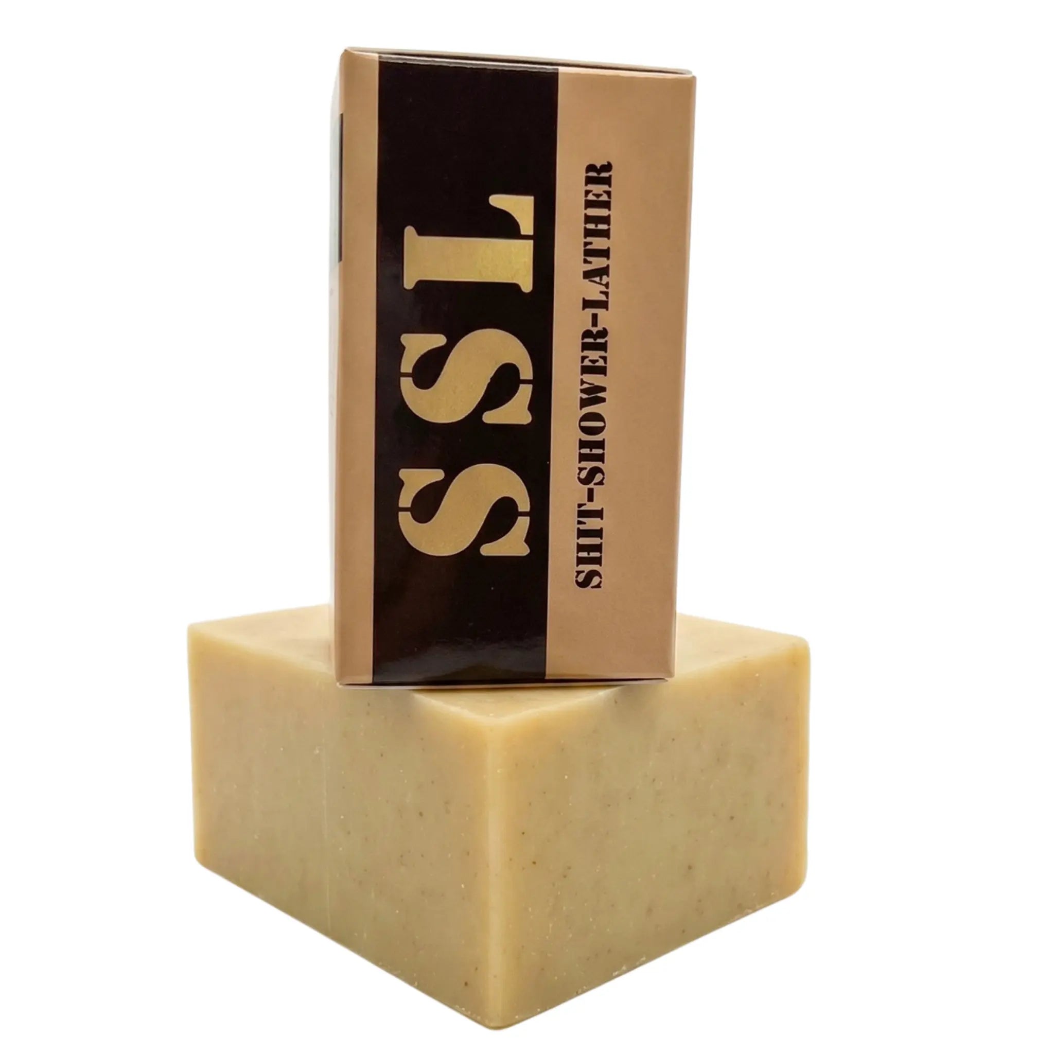 A block of Holy Grail Natural Soap by Flagship Soap Company sits on a white background, featuring a design inspired by military Meals Ready to Eat (MRE) packaging. The tan, textured bar of soap is shown out of the box, with the cardboard packaging balanced on top. The box is tan with a vertical black stripe and bold, stencil-style lettering that reads "SSL" and "SHIT-SHOWER-LATHER," making it a rugged and humorous gift for any veteran.