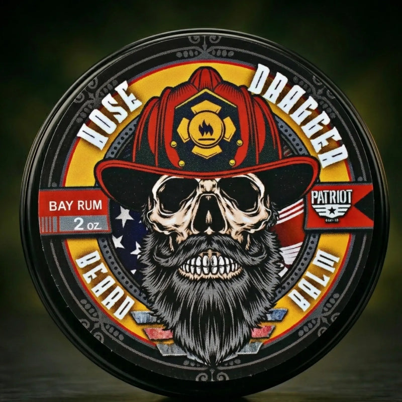 Flagship Soap Co. Hose Dragger Beard Balm (2 oz) in Bay Rum scent. Circular label featuring a bearded skull wearing a red firefighter helmet with a Maltese cross, set against an American flag background. Perfect gift for firefighters and first responders.