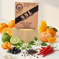 A rugged, military-themed product display for Holy Grail Natural Soap by Flagship Soap Company. The packaging, designed as a "Soap, Ready-to-Lather" (a play on the MRE or Meal, Ready-to-Eat), sits atop a large, creamy bar of soap. The box features a "Shit-Shower-Lather" (SSL) design and notes it is "Warfighter Tested." Surrounding the soap are the fresh scent ingredients: whole oranges, sliced citrus, lime, white blossoms, black peppercorns, and vibrant red peppers. This MRE inspired soap makes a perfect gi