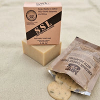 A high-quality bar of Holy Grail Natural Soap by Flagship Soap Company presented in its tan, military-themed packaging, which features a veteran-inspired "SSL" (Shit-Shower-Lather) design. The soap box, marked with a weight of 5.56 OZ, sits atop an actual bar of natural soap. Next to the soap is an open MRE (Meal, Ready-to-Eat) packet of "Cheddar Cheese Sauce with Jalapeños," emphasizing the "Jalapeño and Citrus" scent profile. This ruggedly styled soap makes for a unique gift for those in the military or f