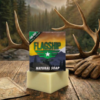 Flagship Soap Company's "The Rut" natural scent control hunting soap bar and box, featuring pine and cedarwood essential oils, displayed in a rustic outdoor setting with elk antlers, a leather glove, and an Idaho map.
