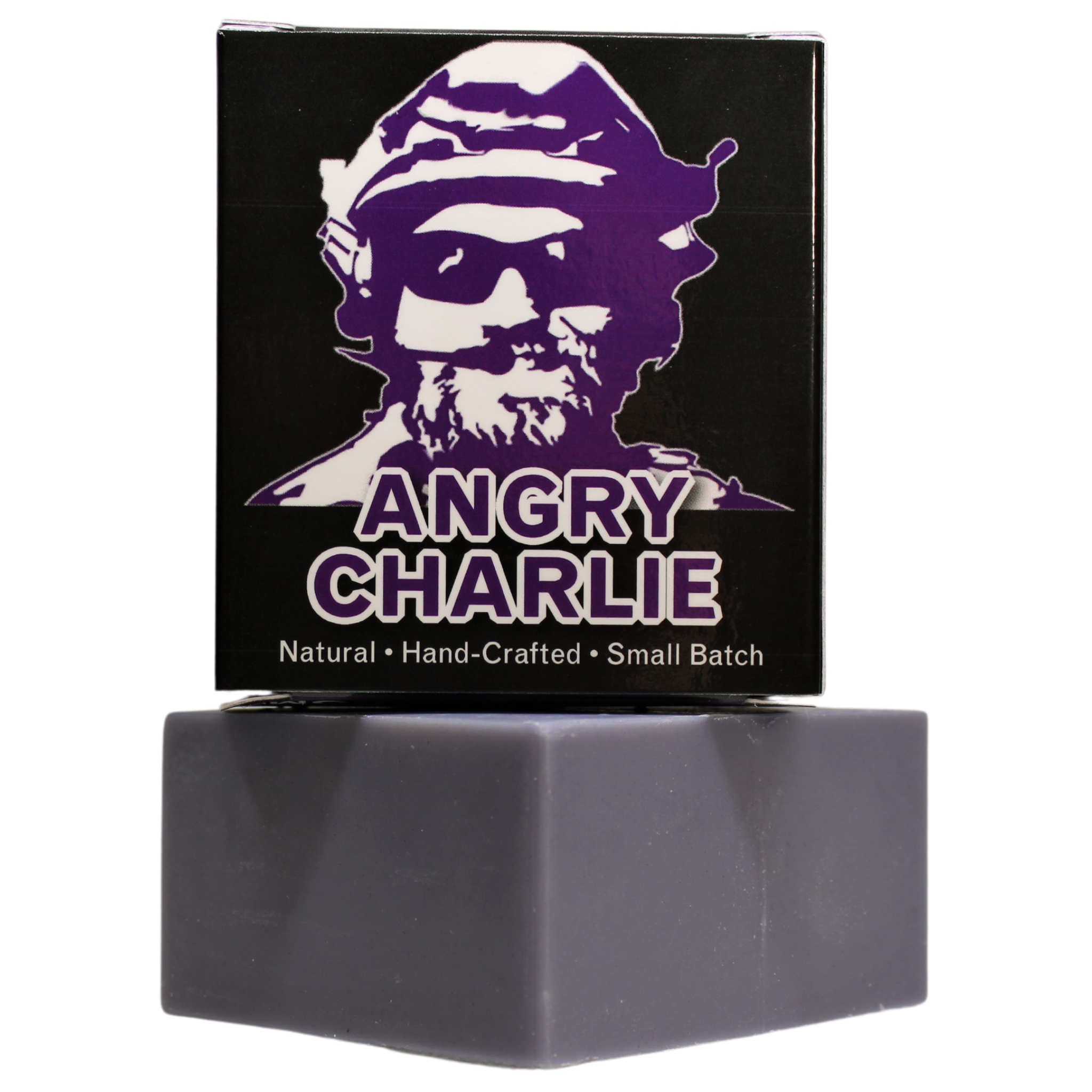 Angry Charlie Natural Soap - Vanilla Cedarwood - Flagship Soap Company - A studio-lit photograph on a white background, featuring a black box of "Angry Charlie" soap stacked on a grey, natural-clay-colored bar of soap. The box has a bold, purple and white portrait of a man in a beret and sunglasses (the "Angry Charlie" figure). The text reads: "ANGRY CHARLIE" in purple and "Natural • Hand-Crafted • Small Batch" in a white footer. The text is crisp and the box is centered over the soap bar.