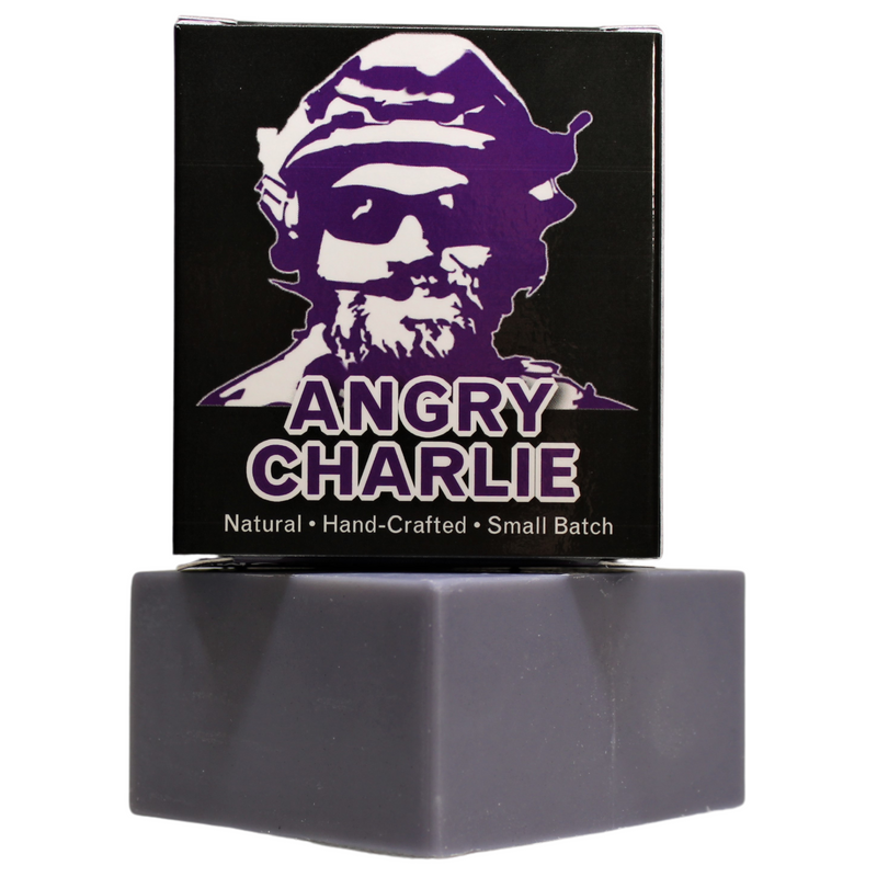 Angry Charlie Natural Soap - Vanilla Cedarwood - Flagship Soap Company - A studio-lit photograph on a white background, featuring a black box of "Angry Charlie" soap stacked on a grey, natural-clay-colored bar of soap. The box has a bold, purple and white portrait of a man in a beret and sunglasses (the "Angry Charlie" figure). The text reads: "ANGRY CHARLIE" in purple and "Natural • Hand-Crafted • Small Batch" in a white footer. The text is crisp and the box is centered over the soap bar.